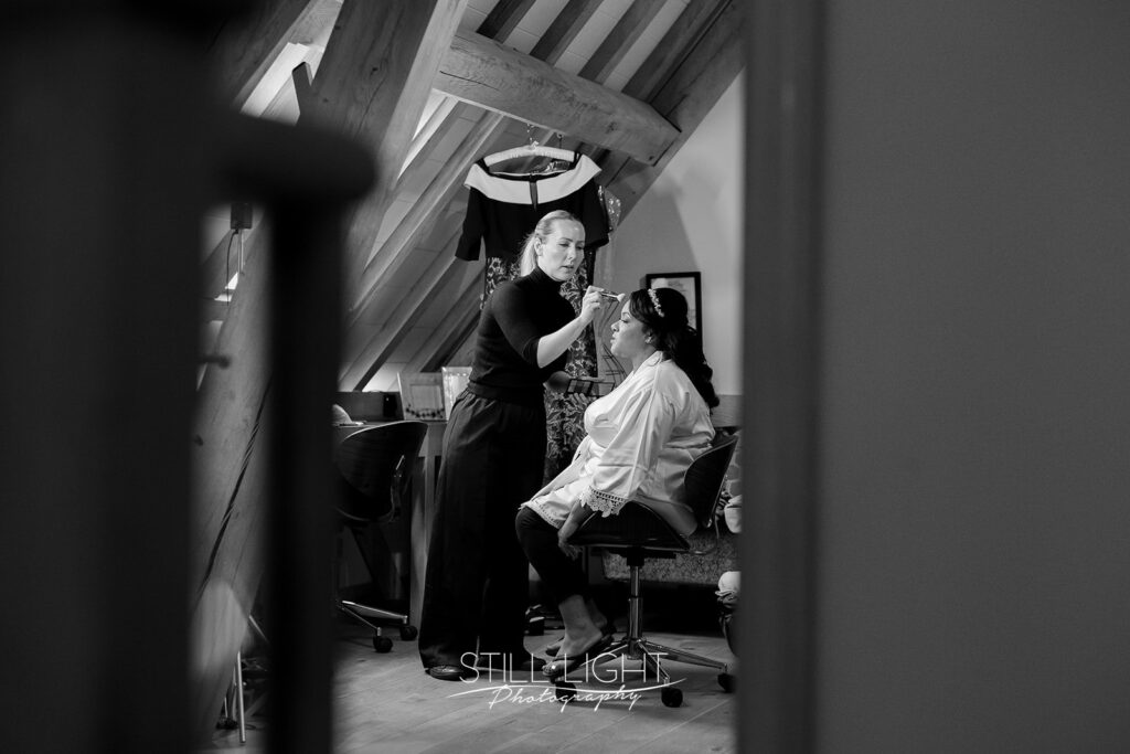 black and white image of bride having her amkeup done shot through doorway at redhouse barn during bridal preparation