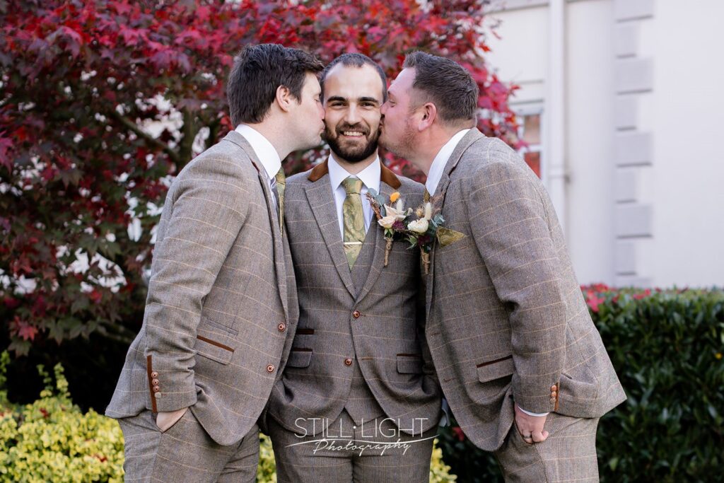 groom being kissed on the cheek by both best men at brockencote hall