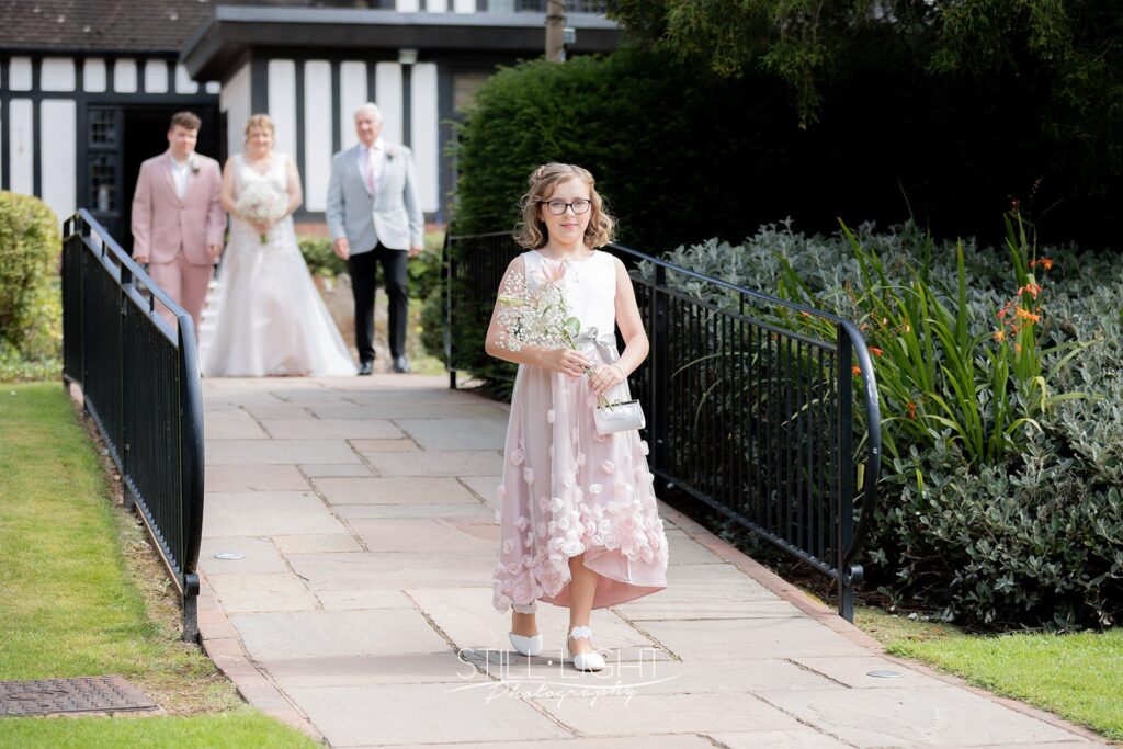 fowergirl Lily walking towards wedding ceremony holding a bunch of lilies with bride behind her