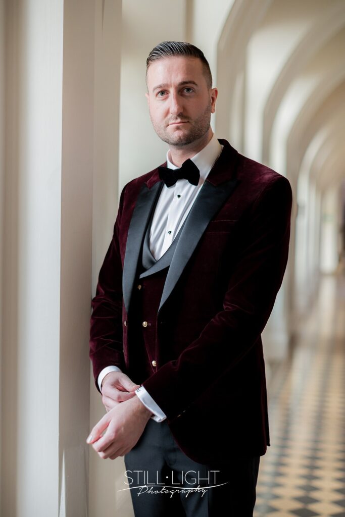 groom in the cloisters of stanbrook abbey hotel wearing red velvet jacket looking at camera