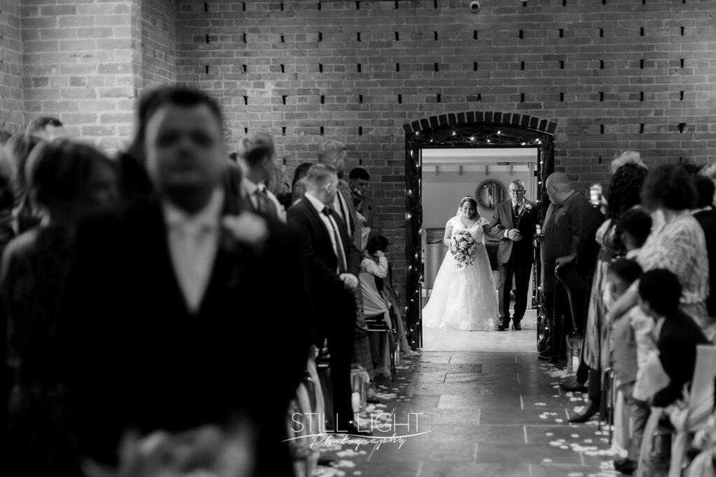 black and white image of bride and her father in doorway getting ready to walk into red brick barn for wedding ceremony