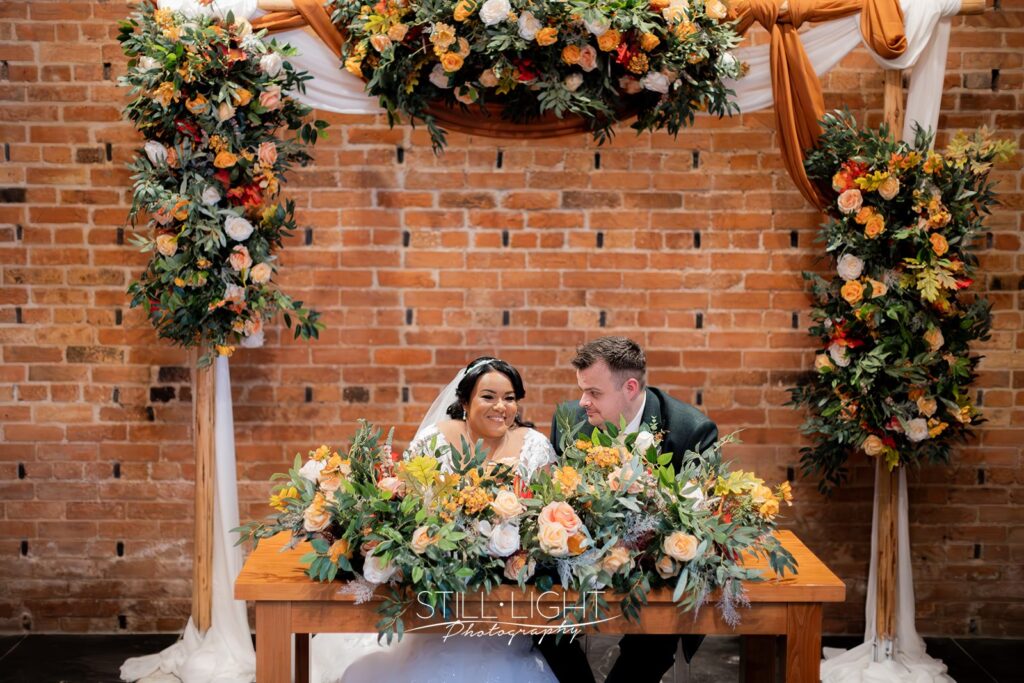 bride and groom sat at table behind lots of autumn flowers signing the register on their wedding day at redhouse barn