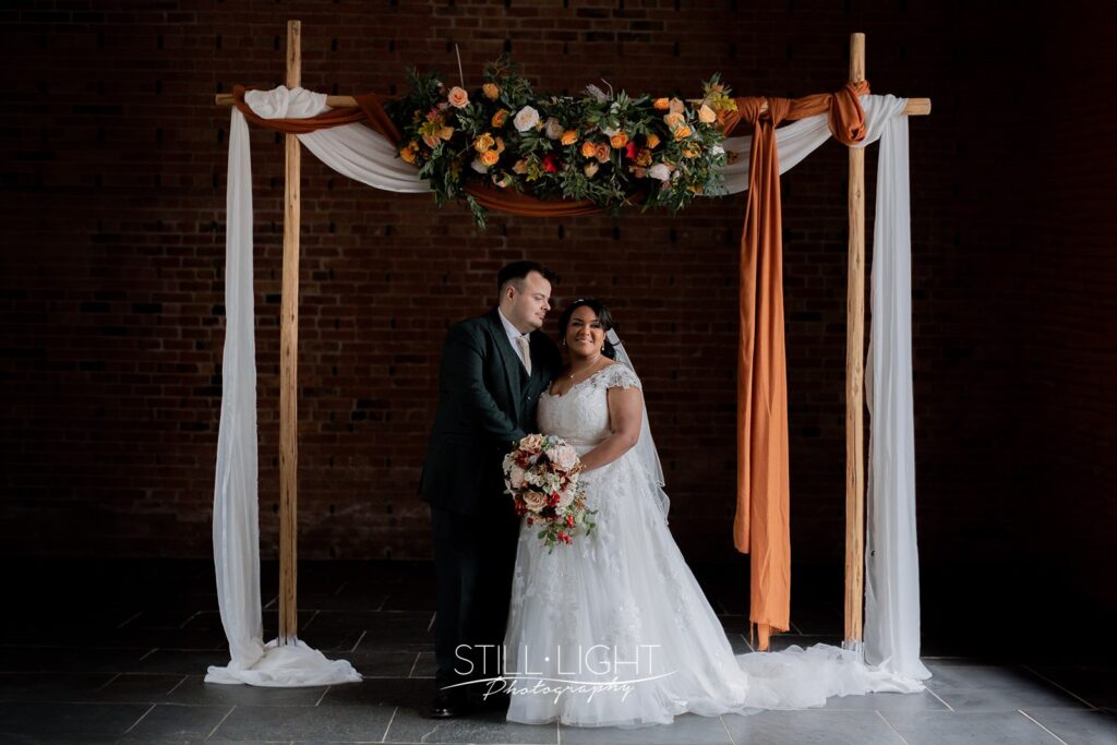 bride and groom standing together under wooden arch with autumn flowers and drapes