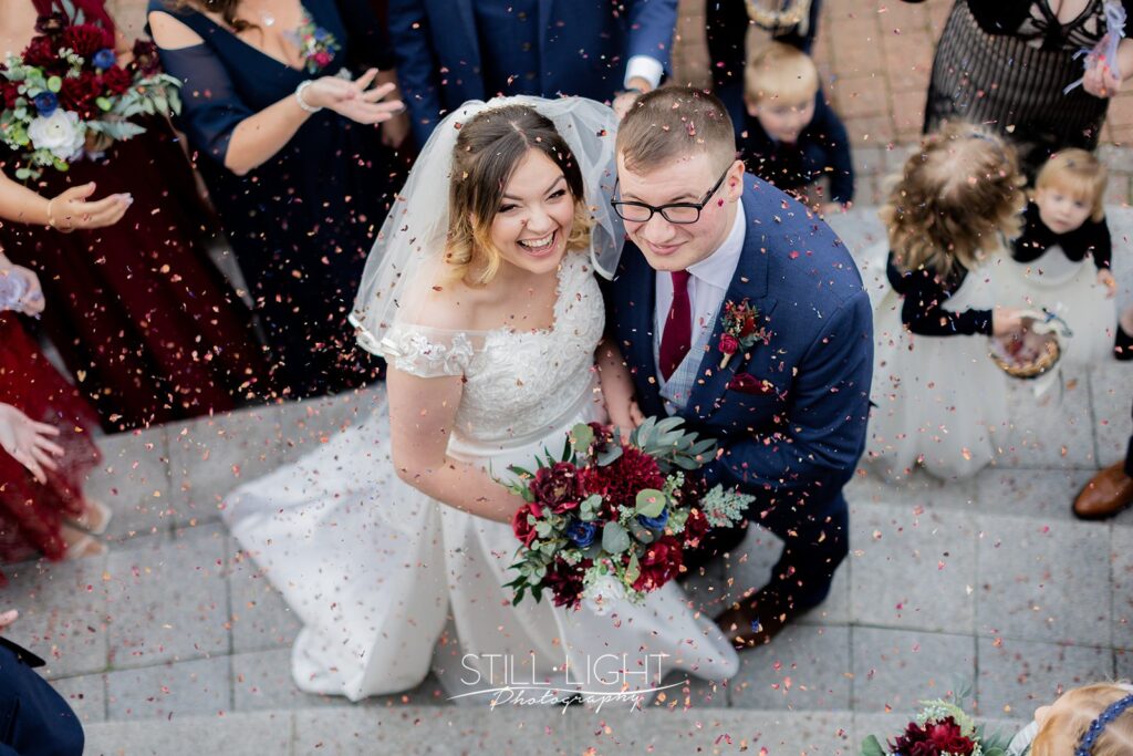 bride and groom looking up at camera laughing as confetti is thrown from balcony at wootton park