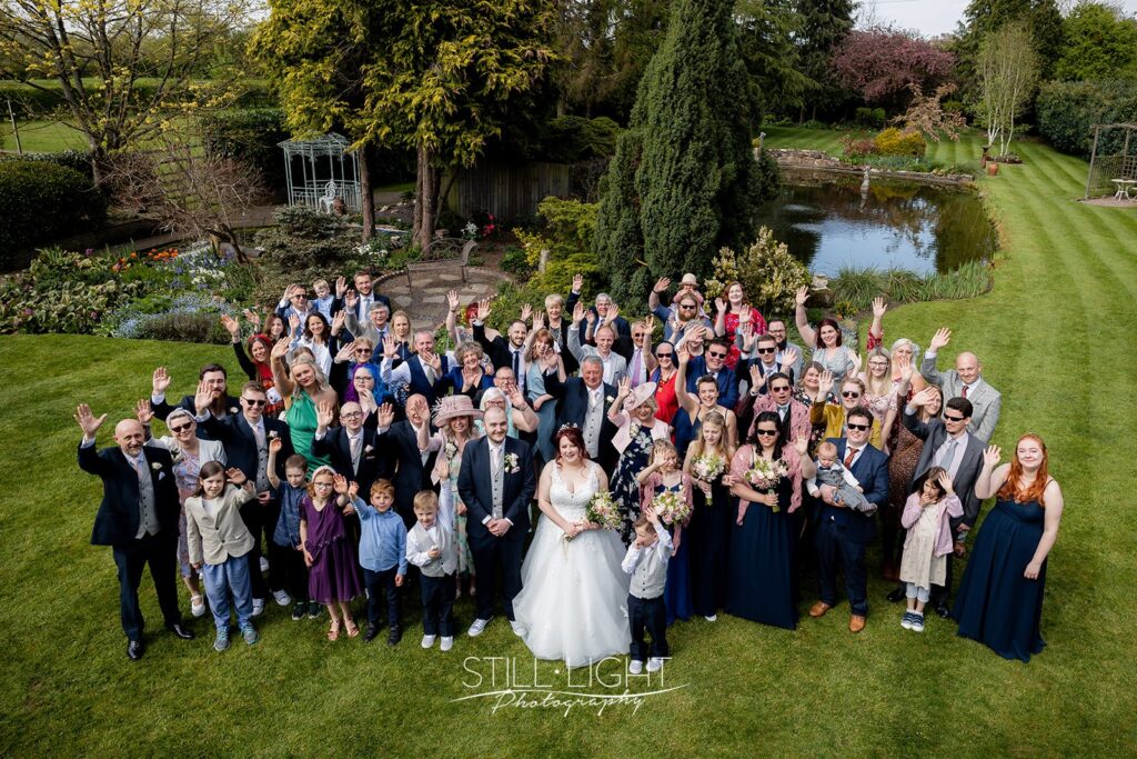group photograph taken from balcony during weding at redhouse barn with gardens and pond in background