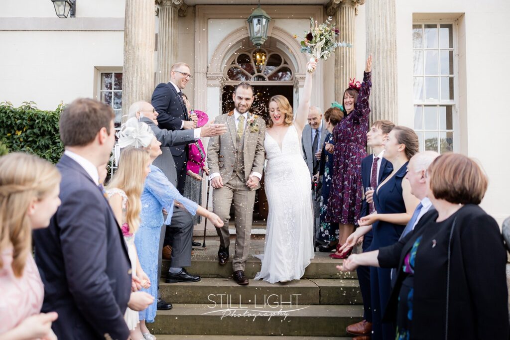 bride and groom cheering as they walk down steps of brockencote hall to guests throwing confetti
