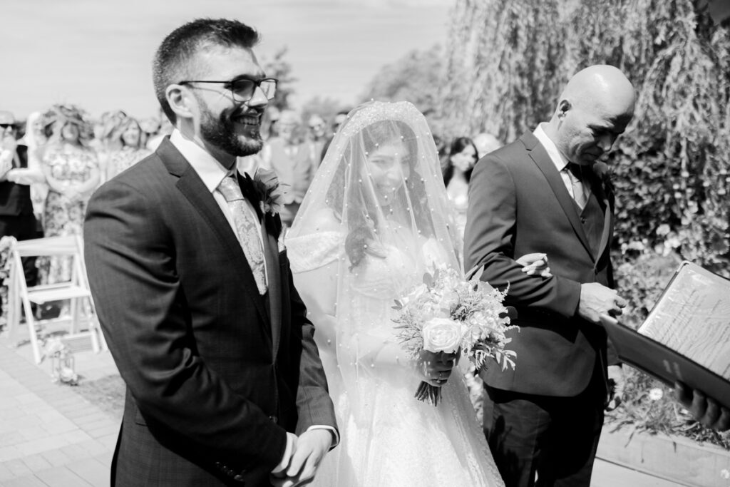 black and white image of bride wearing veil over her face as she meets the groom during their lakeside wedding ceremony