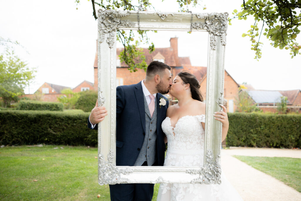bride and groom kissing behind photo frame hanging on tree during drinks reception at bordesley park