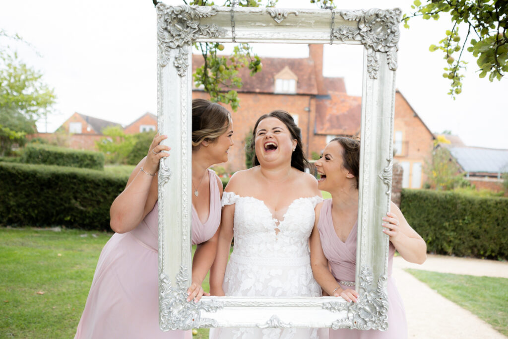 bride and bridesmaids laughing behind photo frame during drinks reception at bordesley park