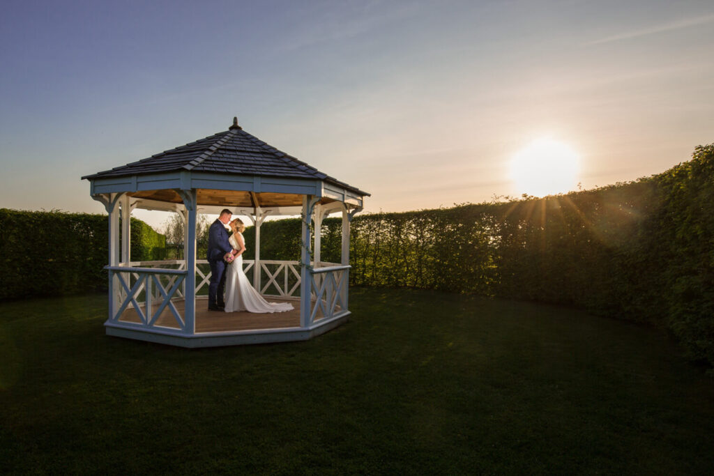 couple standing inside bandstand as the sun drops behind a hedge, taken by experienced wedding photographers still light photography