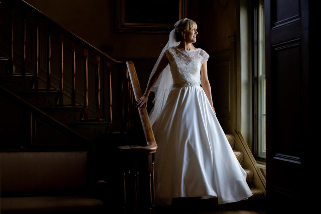 bride standing on staircase looking out of window with beautiful natural light by experienced wedding photographers still light photography