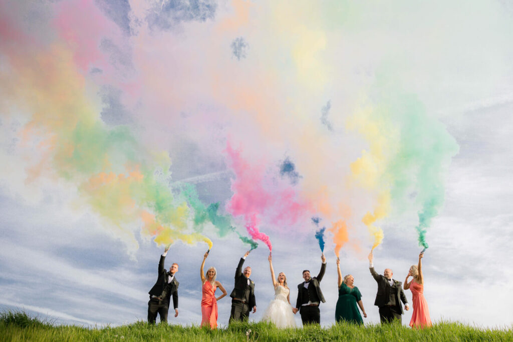 bride and groom and their friends holding up colourful smoke grenades against blue hazy sky