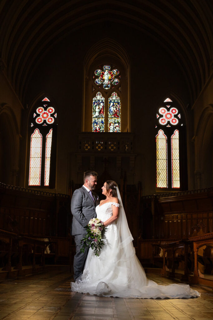 couple on their wedding day standing against dark background and stained glass windows at stanbrook abbey