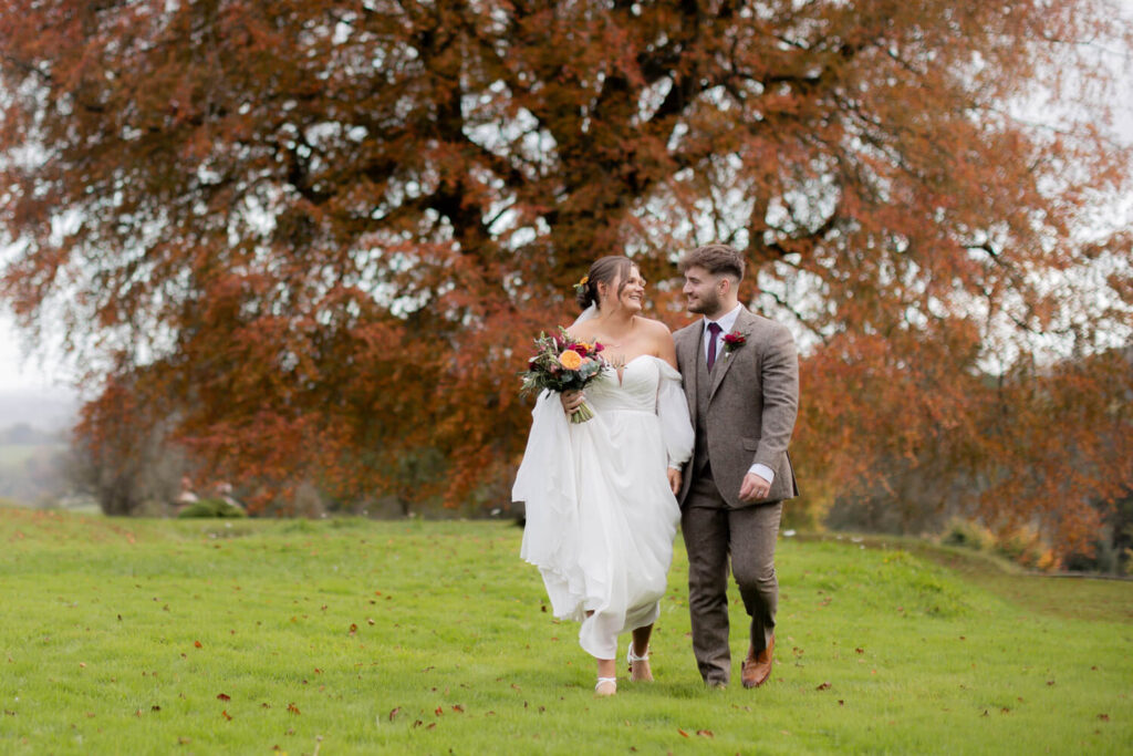 couple walking towards camera chatting backdrop of autumnal oak tree at bredenbury court barns
