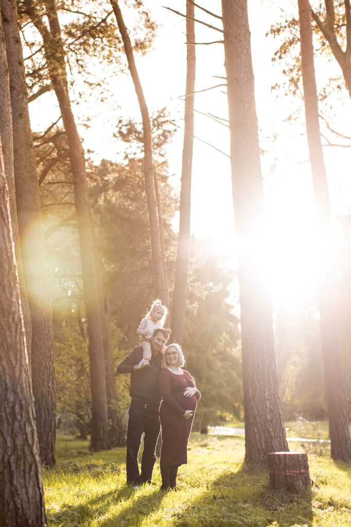 couple with young daughter on shoulders standing in trees with sunshine haze  during an outdoor family photography session in worcestershire