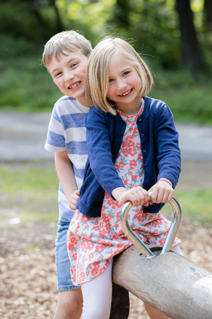 siblings boy and girl sitting together smiling at camera on a seesaw during an outdoor family photography session in worcestershire