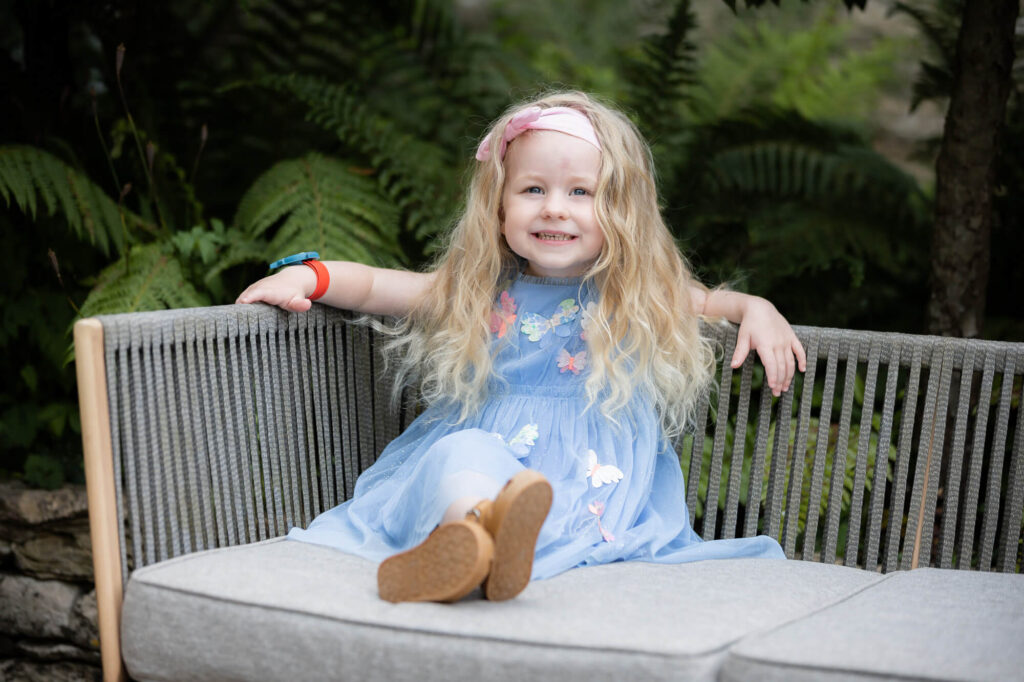 little girl with blond hair in blue dress smiling at camera sat on bench during an outdoor family photography session in worcestershire