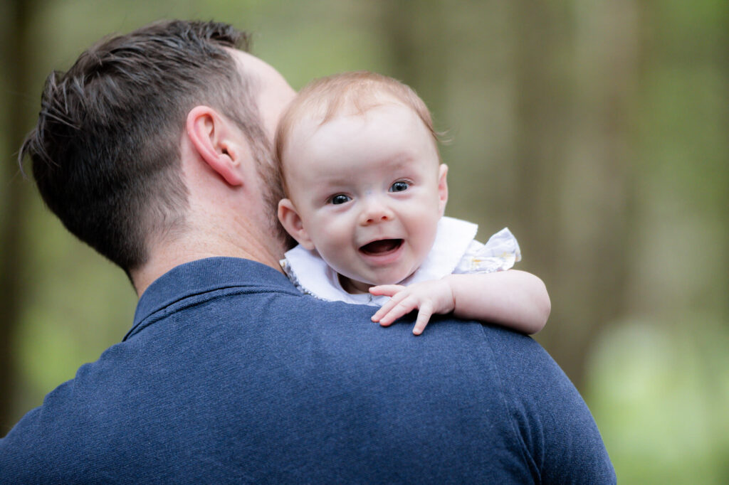 baby over dad's shoulder smiling at camera  during an outdoor family photography session in worcestershire