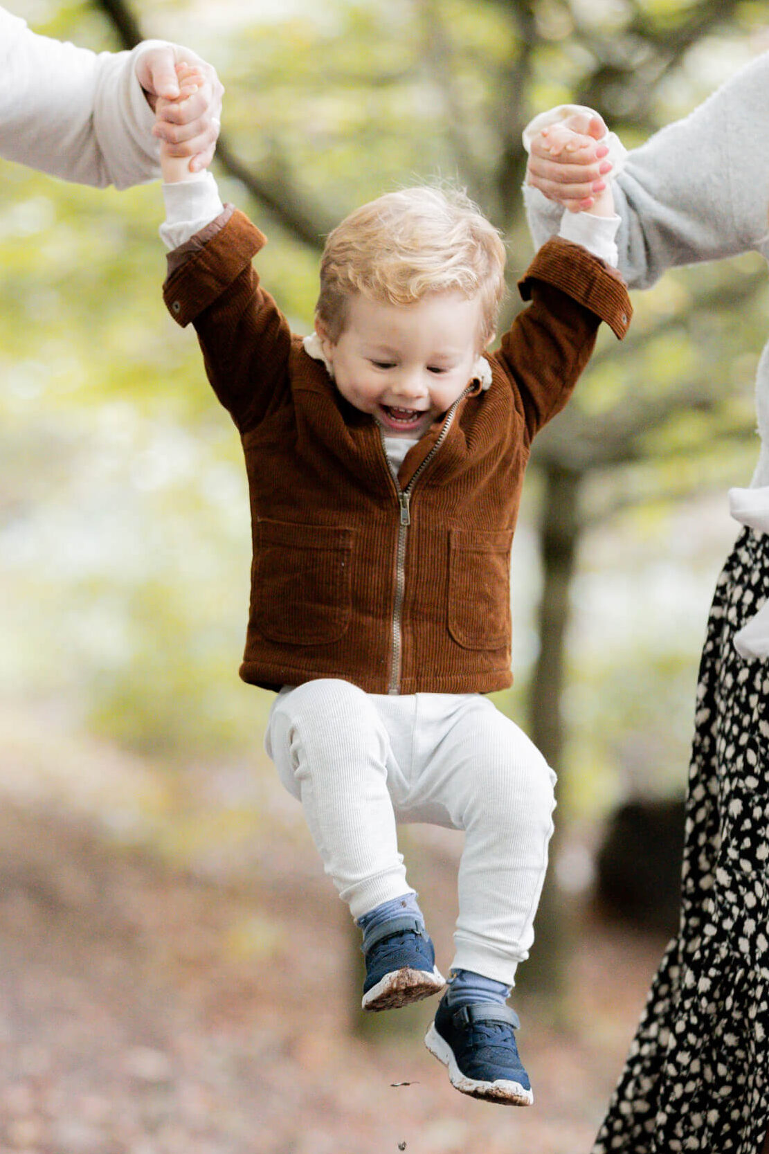 little boy wearing brown coat laughing as he is being swung in parents hands  during an outdoor family photography session in worcestershire