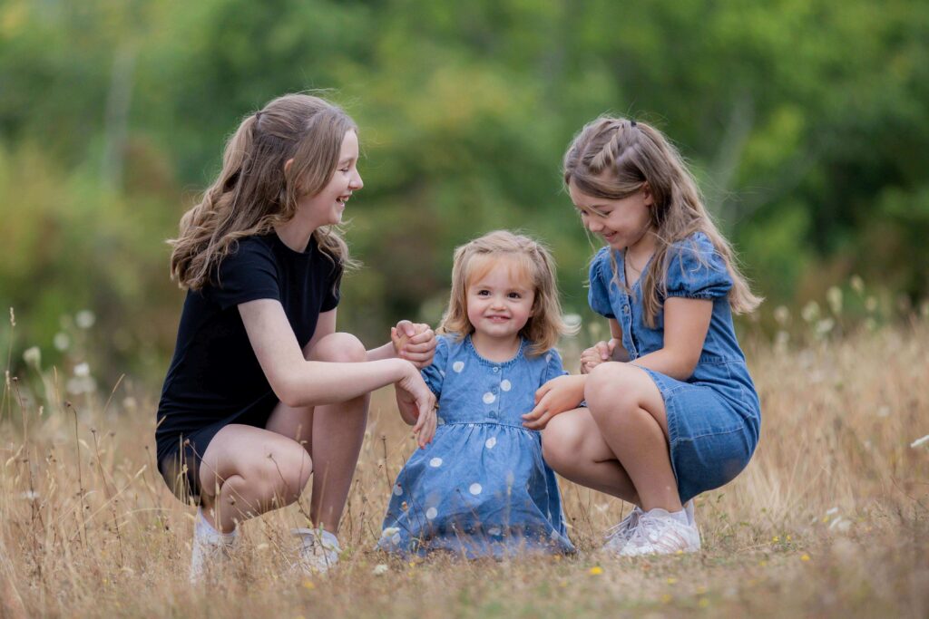 three sisters young girls crouching together in field smiling at each other  during an outdoor family photography session in worcestershire