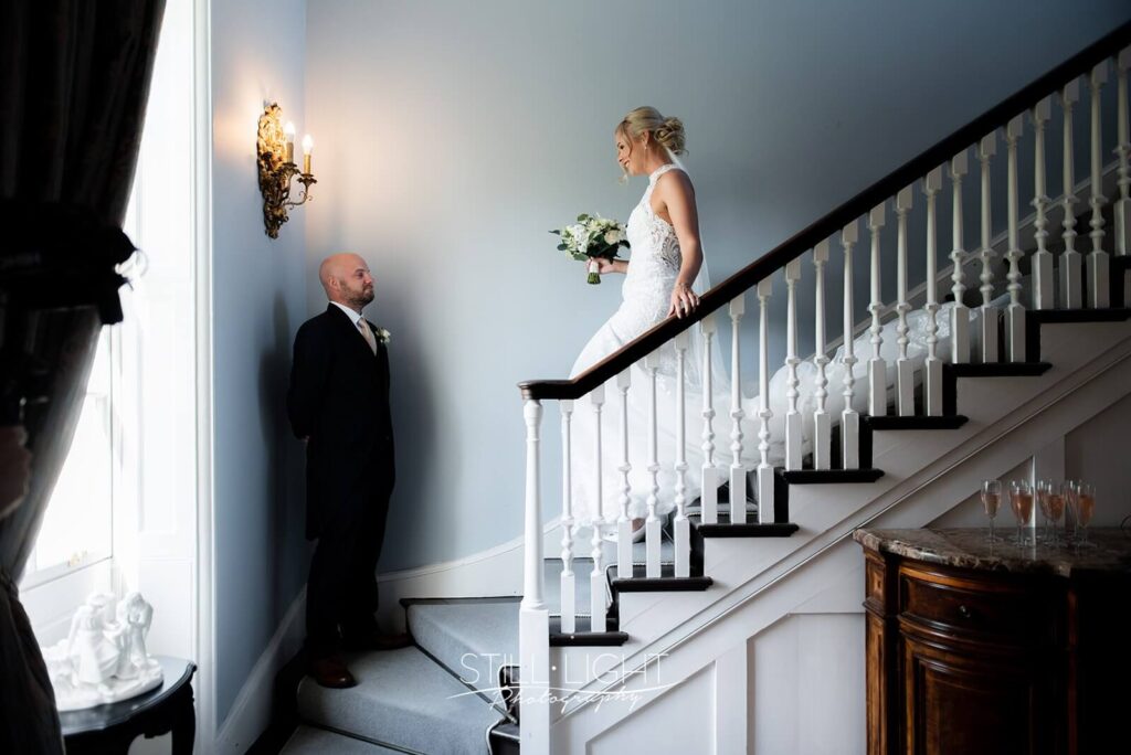 bride walking down stairs towards her brother holding white bouquet in brides manor at stanbrook abbey