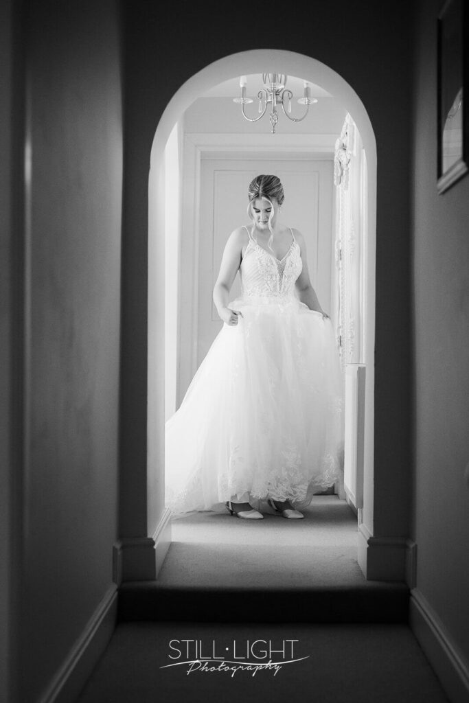 black and white image of bride adjusting wedding dress standing in corridor in brides manor stanbrook abbey