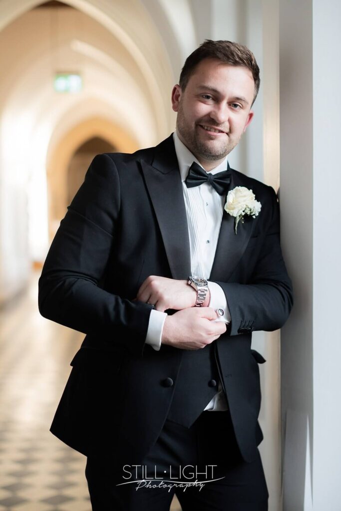 groom poses for camera whilst leaning on wall indoor in cloisters at stanbrook abbey