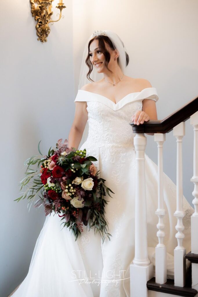 bride standing on staircase holding large red bouquet in brides manor at stanbrook abbey