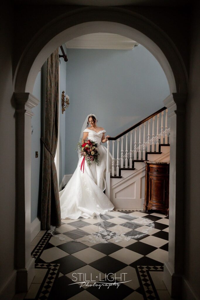taken throguh archway of bride standing on staircase holding large red bouquet in brides manor at stanbrook abbey