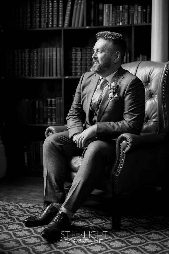 black and white photo of man sitting in wingback armchair looking out of the window in library
