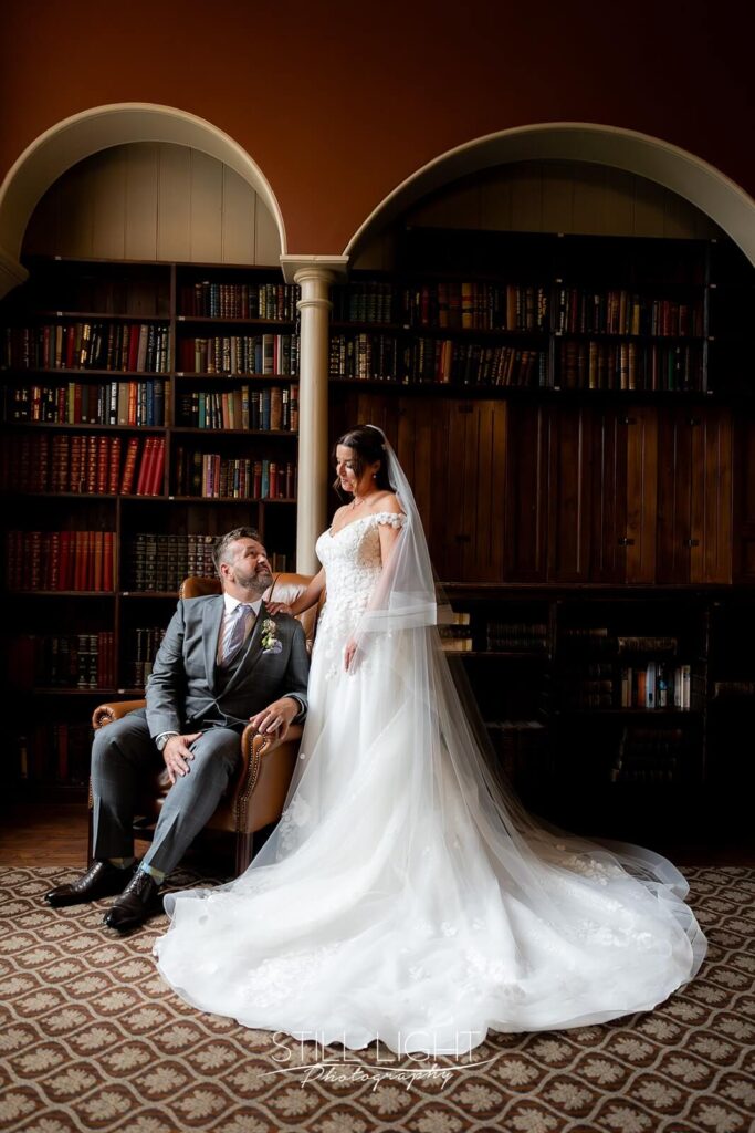 bride and groom pose for photo with groom sitting and bride standing in library bar at stanbrook abbey
