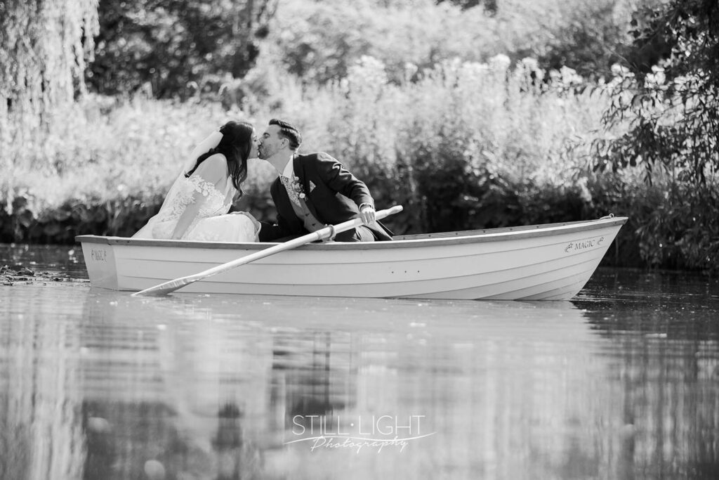 black and white image of couple sitting in rowing boat kissing on wedding day at wootton park