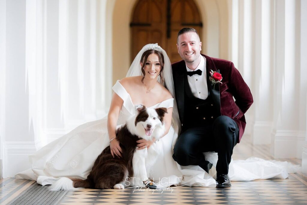 bride and groom crouched with their dog posing for camera indoor in cloisters at stanbrook abbey