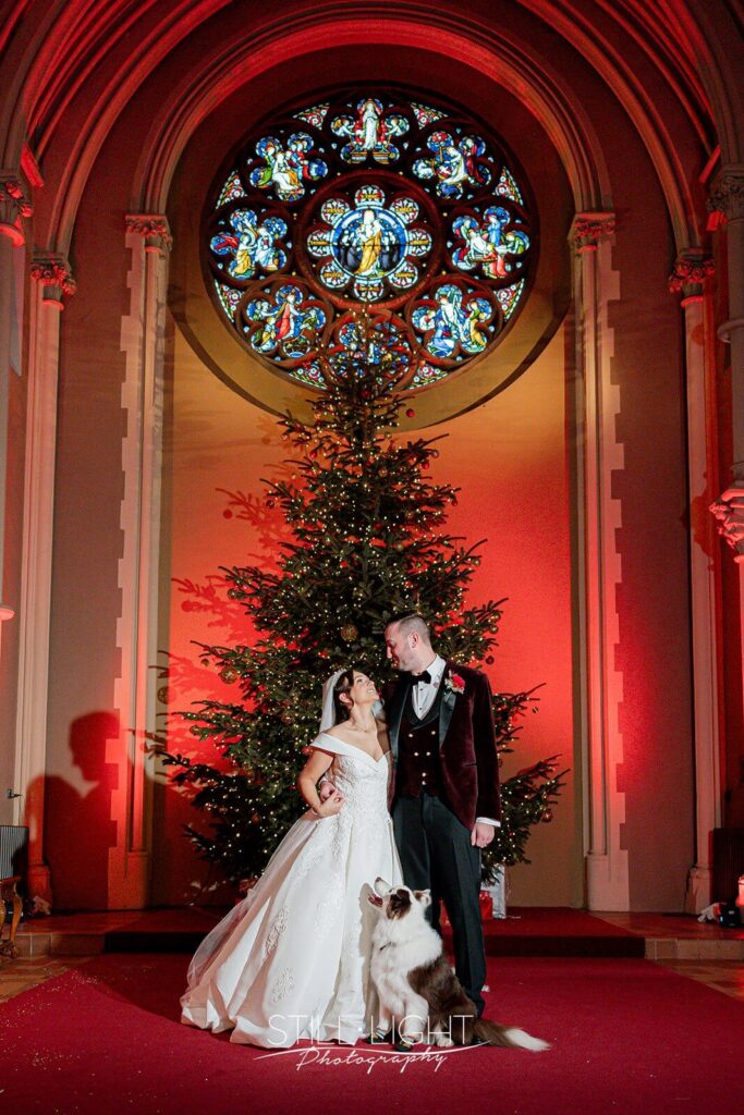 bride and groom standing in front of christmas tree with their dog looking up at them from their feet Callow Great Hall at Stanbrook Abbey