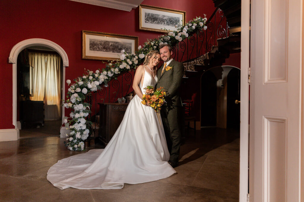 bride and groom standing at the foot of the staircase at hopton court on wedding day holding yellow and orange bouquet

