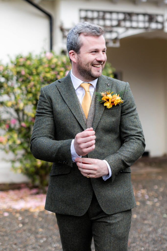 man walks towards camera adjusting his sleeve cuff of a green tweed suit at hopton court wedding
