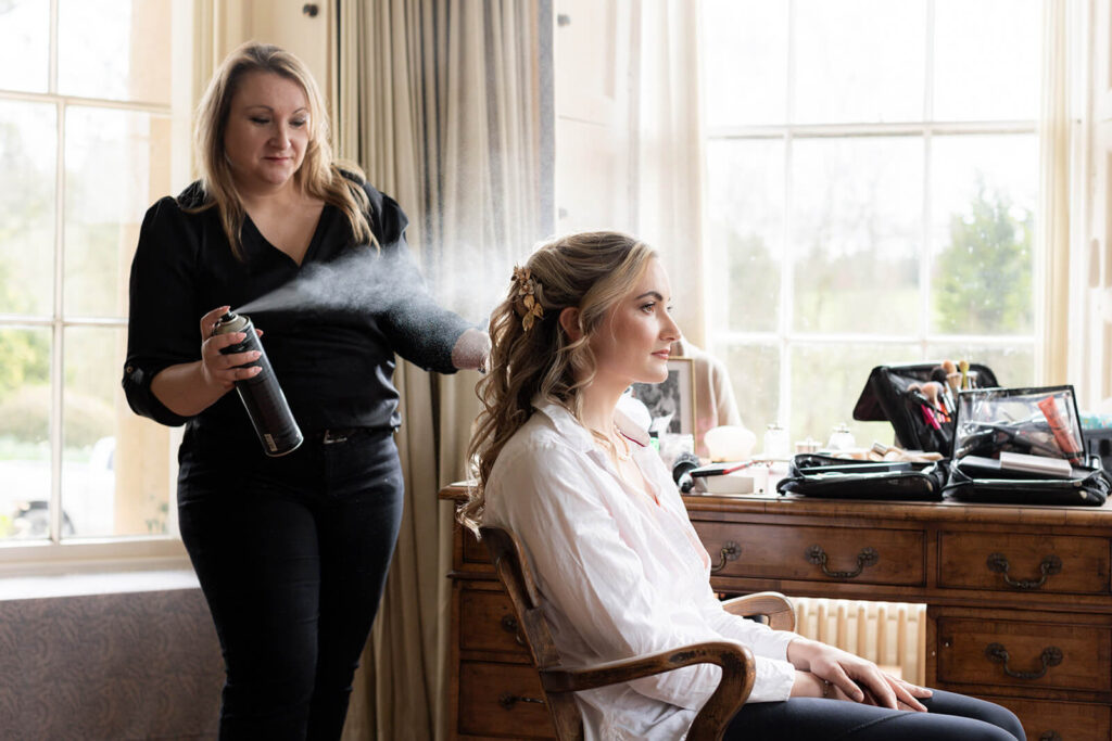 hairspray being sprayed onto brides hair as she sits in chair in the bay windows of the house at hopton court wedding
