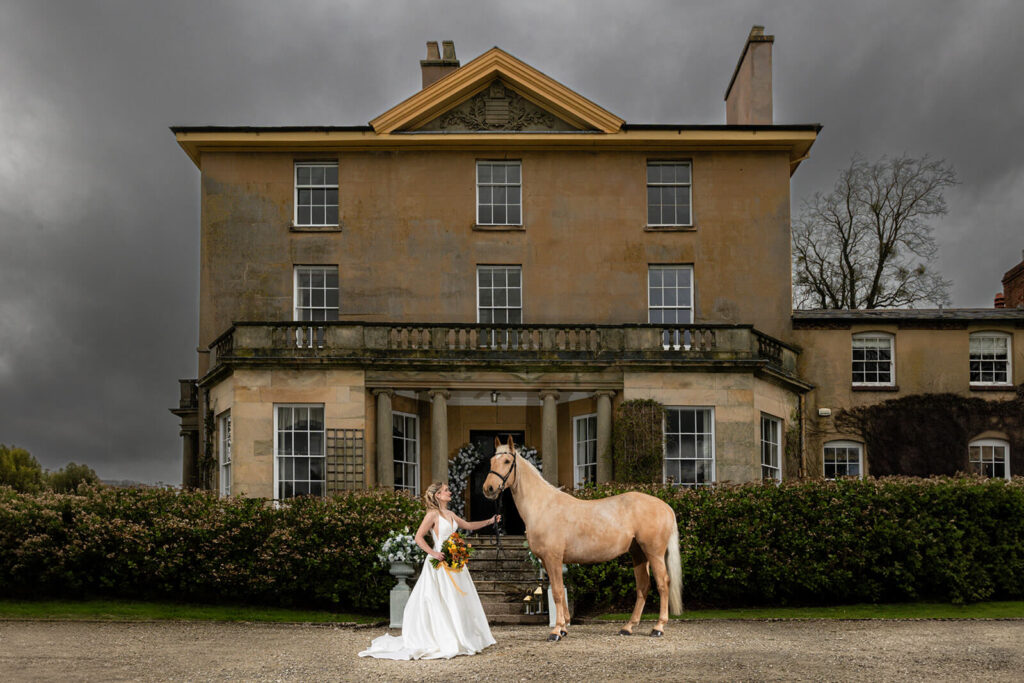 bride stands with palomin horse in front of Georgian Manor at Hopton Court on wedding day