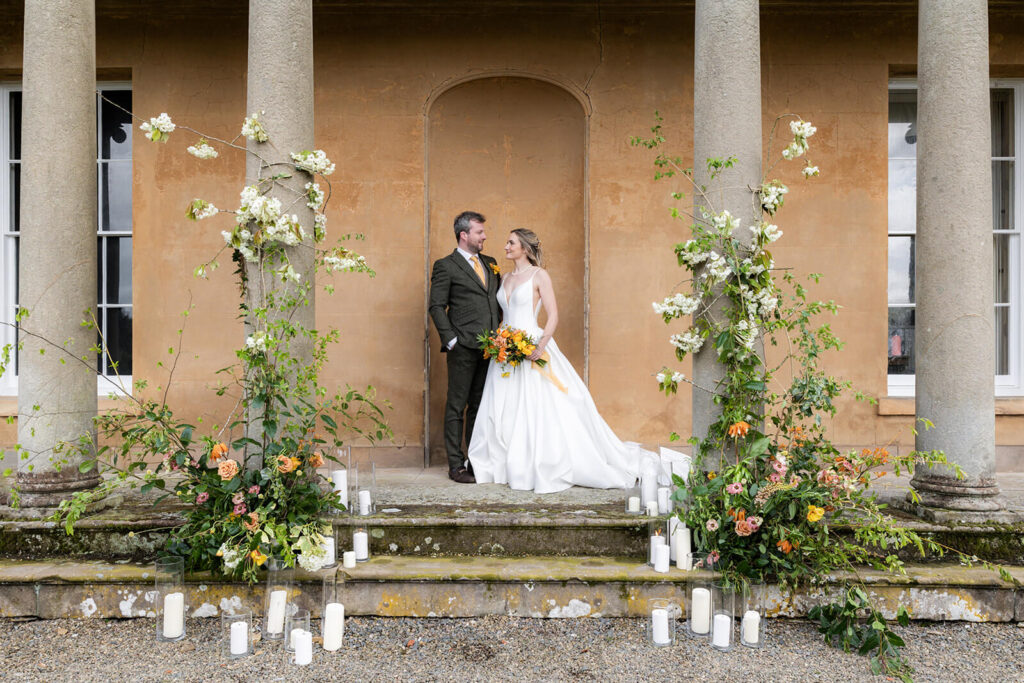 bride and groom standing rogehter on steps of hopton court in between the pillars surrounded by rustic floral displays and white candles