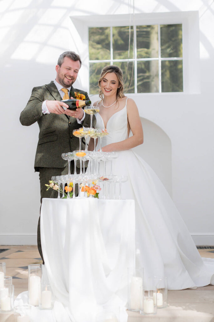 bride and groom pour champagne into champagne glass tower at hopton court wedding
