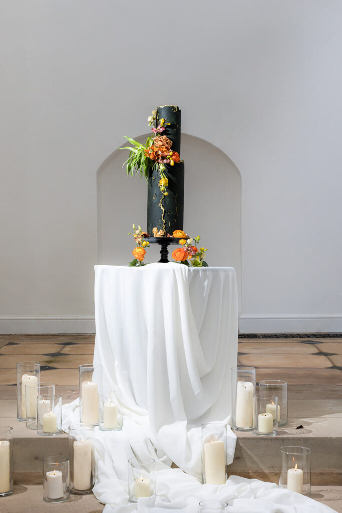 dark green tall narrow wedding cake with yellow and orange flowers on white table surrounded by candles