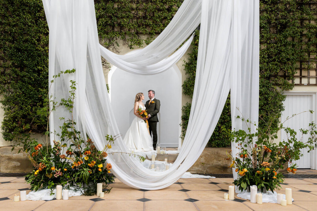 bride and groom stand together behind white drapes and beautiful orange and green floral displays in the orangery at hopton court wedding
