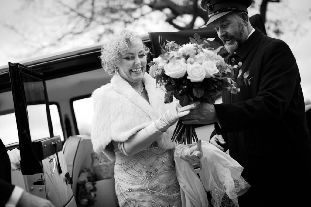 black and white image of smiling bride getting out of car and being handed her bouquet