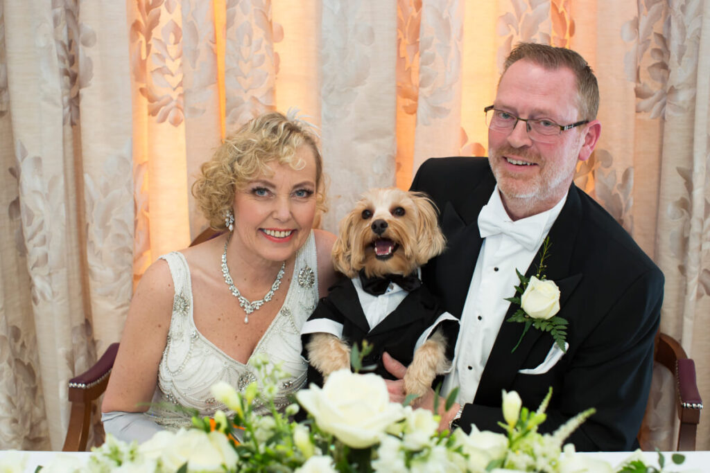 bride and groom sitting at table with flowers and their dog in between them on wedding day at Brockencote Hall, a dog friendly wedding venue