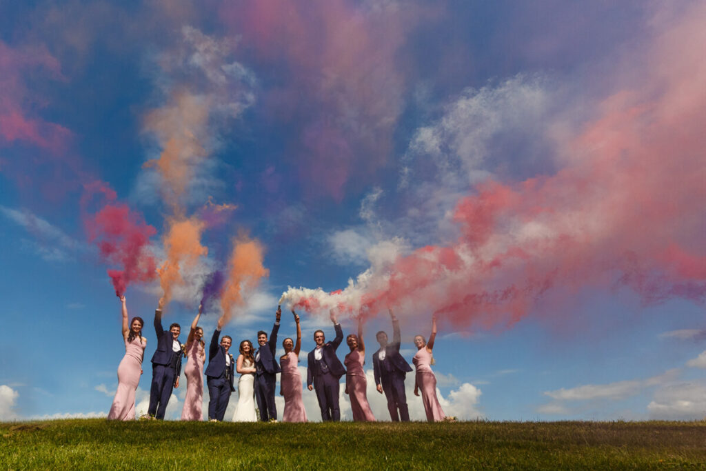 bride groom and wedding party holding colourful smoke grenades up towards bright blue sky