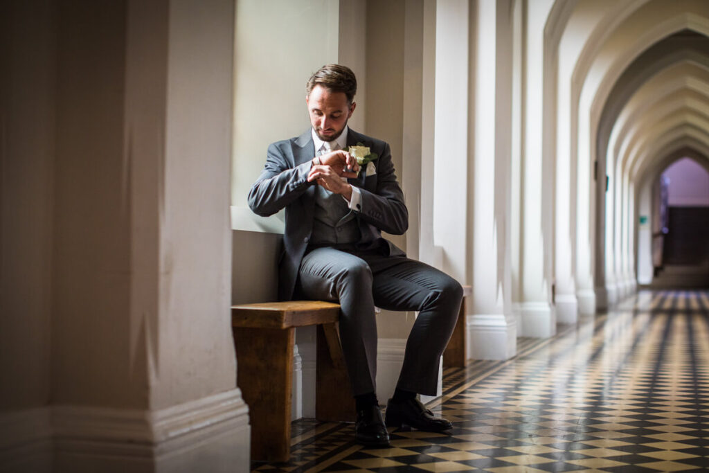 groom sitting on bench adjusting his cufflings indoor in cloisters at stanbrook abbey