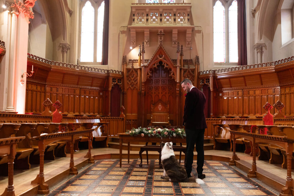 groom stood at front of callow great hall in stanbrook abbey waiting for his bride with his dog sitting by his side